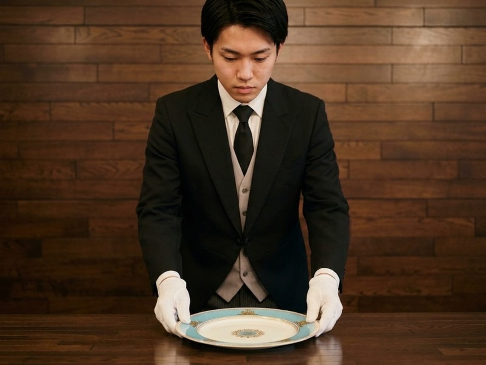日本執事アカデミーの公式サイトより。モーニングコートと白手袋の若い執事が金彩の皿を両手で捧げ持つ場面